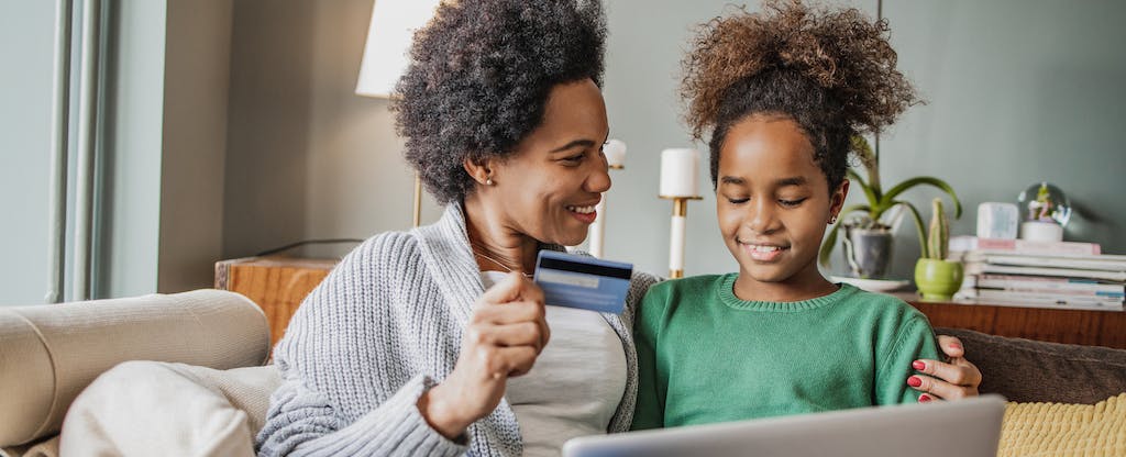 A smiling parent sitting on a couch next to their child, holding a laptop and showing them how to use their Greenligh debit card